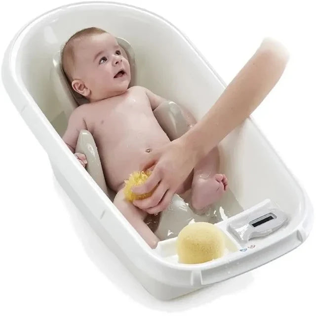 A baby sitting in a white plastic bath tub while an adult hand washes the baby's legs with a yellow sponge. A small digital thermometer is visible on the front right of the tub.