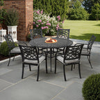 A 7-piece outdoor patio dining set featuring a round black metal table and four matching armchairs with light gray cushions, arranged on a gray stone patio in a landscaped backyard.