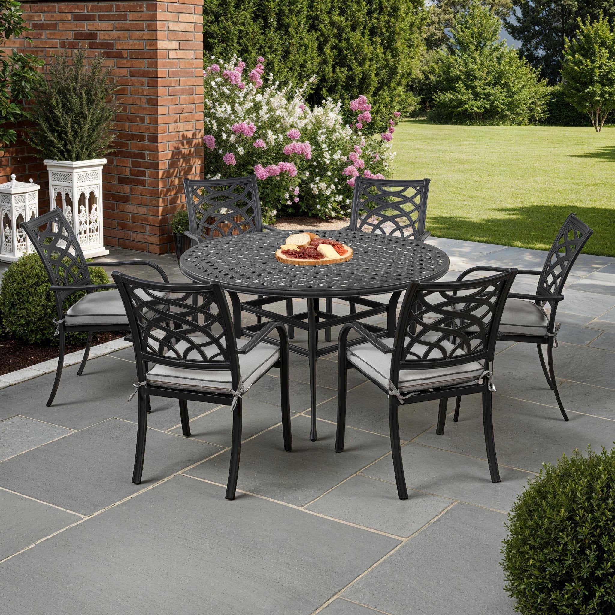 A 7-piece outdoor patio dining set featuring a round black metal table and four matching armchairs with light gray cushions, arranged on a gray stone patio in a landscaped backyard.
